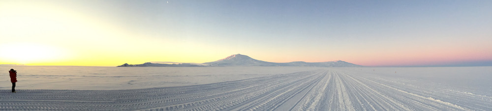 A view of Ross Island at sunset. Mark is standing at the left side of this image and the sun is setting behind the Transantarctic Mountains. Ross Island, with Mt. Erebus, is in the center of the photograph. On the right side of the image you can see the blue shadow of the Earth above the horizon and the pink glow of the sun still illuminating the higher levels of the atmosphere. A thin crescent moon is visible at the top of the image about a third of the way from the left side of the photograph. 