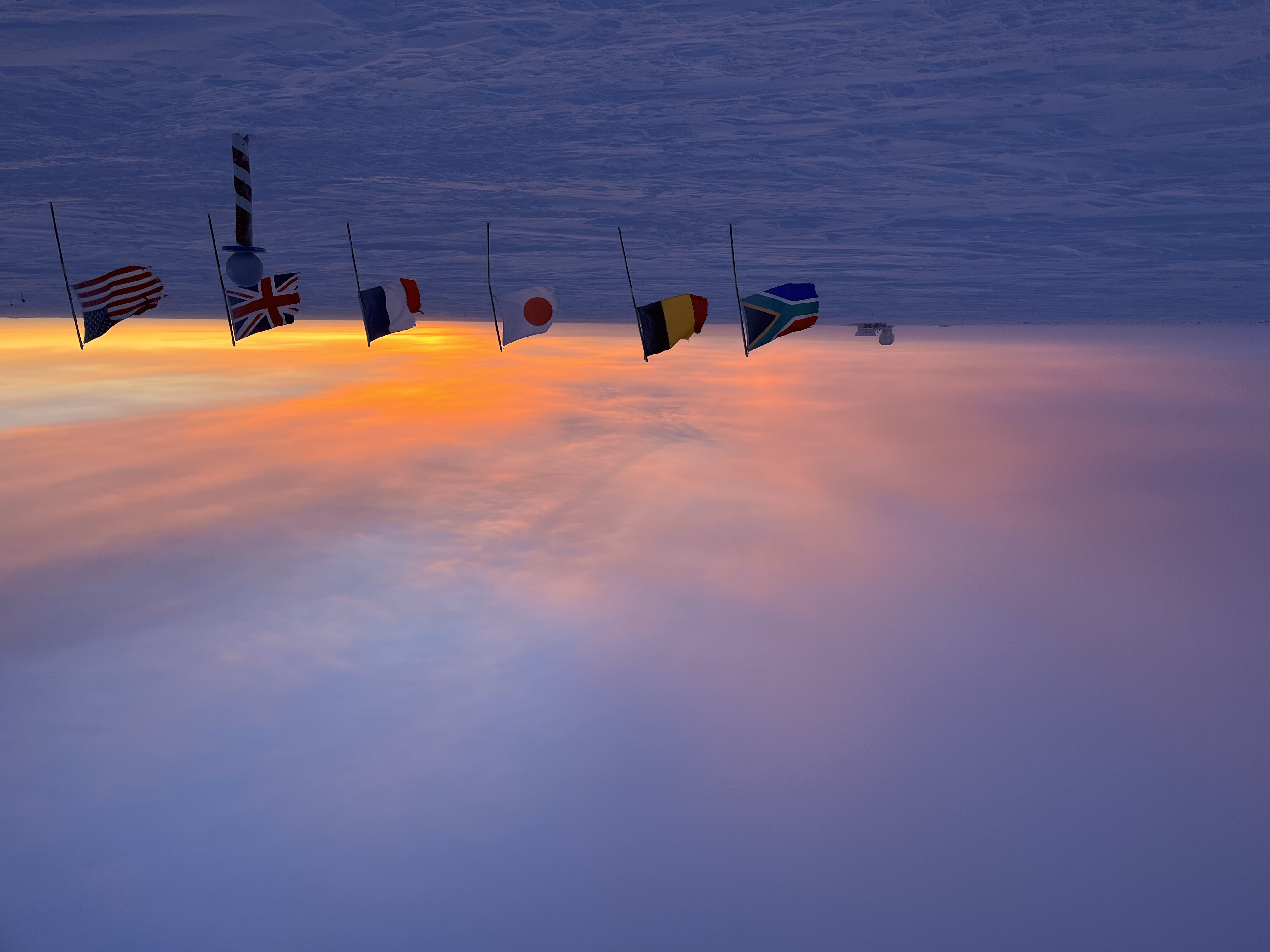 Faint light from the sun is visible on the horizon at the South Pole, across a vast stretch of snow with a row of flags across the forefront.